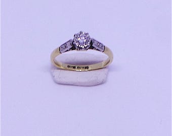 May include: A gold and silver ring with a central cluster of small diamonds. The ring has a gold band and silver accents on the shoulders, with small diamonds set into the design. The ring is displayed on a white surface.