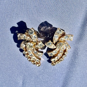 Puede incluir: Un par de pendientes vintage dorados con clip. Cada pendiente presenta un diseño en espiral en forma de abanico incrustado con brillantes pedrería transparente. Los pendientes están colocados sobre un fondo de tela azul claro.