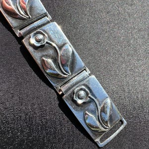 May include: A silver bracelet with a floral design. Each rectangular link features a raised flower and leaf motif. The bracelet is connected by hinged links and has a clasp closure. The metal has a polished finish.