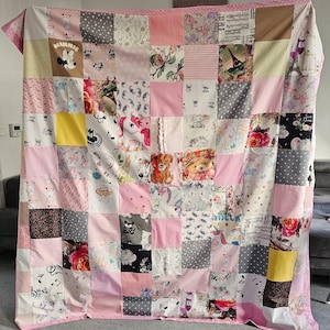 Op de afbeelding: Een patchwork quilt met verschillende stoffen in roze, wit, grijs en bruin. De quilt bevat vierkanten met bloempatronen, stippen, muzieknoten en stripfiguren zoals Minnie Mouse en eenhoorns. De quilt heeft een roze rand.