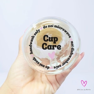 May include: A clear glass cup with a pink heart and the text "Cup Care" in black. The text around the cup reads "handwash only do not microwave do not soak not dishwasher safe".