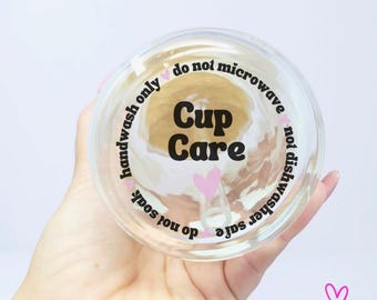 Puede incluir: Una taza de vidrio transparente con un corazón rosa y el texto "Cup Care" en negro. El texto alrededor de la taza dice "handwash only do not microwave do not soak not dishwasher safe".