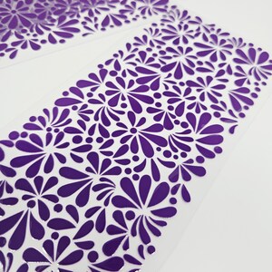 May include: Close-up of two sheets of purple floral patterned material. The design features stylized flowers and teardrop shapes in a repeating pattern. The material appears to be translucent with a glossy finish, set against a white background.