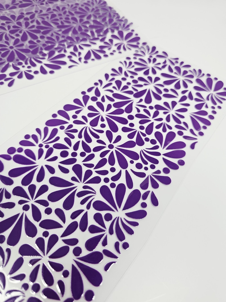 May include: Close-up of two sheets of purple floral patterned material. The design features stylized flowers and teardrop shapes in a repeating pattern. The material appears to be translucent with a glossy finish, set against a white background.
