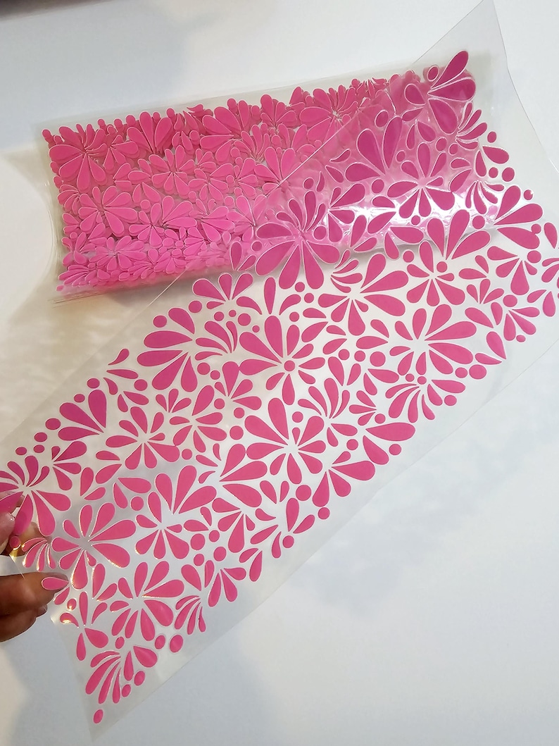 May include: A clear plastic sheet with a pink floral pattern. The sheet is filled with pink flower-shaped cutouts. The design features various sizes of stylized flowers and petal shapes, creating a decorative element.