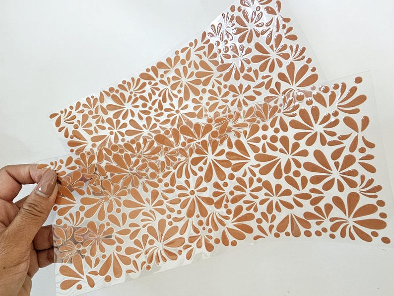 May include: Two sheets of clear, flexible material with a repeating floral pattern in a warm, earthy orange color. The design features stylized flowers and droplet shapes, creating a textured, decorative effect.