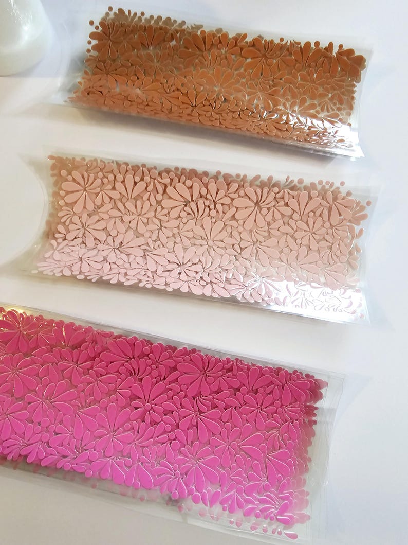 May include: Three pillow-shaped packages filled with floral-shaped embellishments. The top package is filled with brown embellishments, the middle with pink, and the bottom with hot pink. The embellishments are in clear plastic packaging.