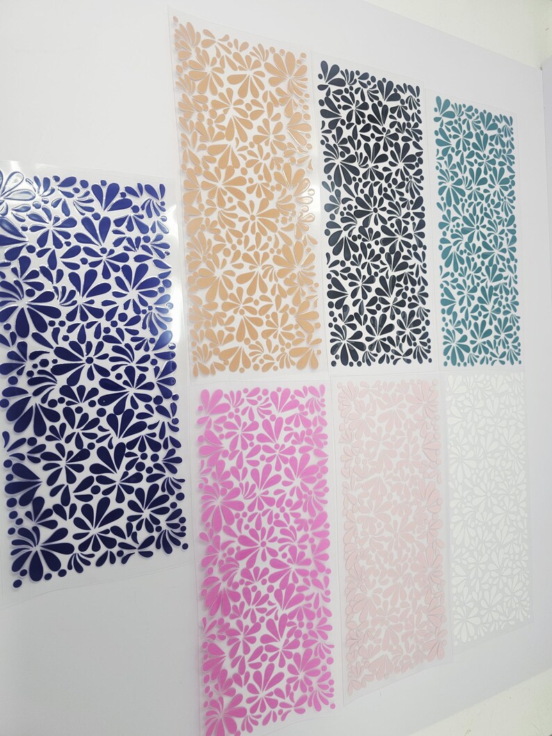May include: A collection of floral pattern sheets in various colors, including navy blue, tan, black, teal, pink, light pink, and white. The sheets are arranged vertically, showcasing the repeating flower design. The background is a plain white surface.