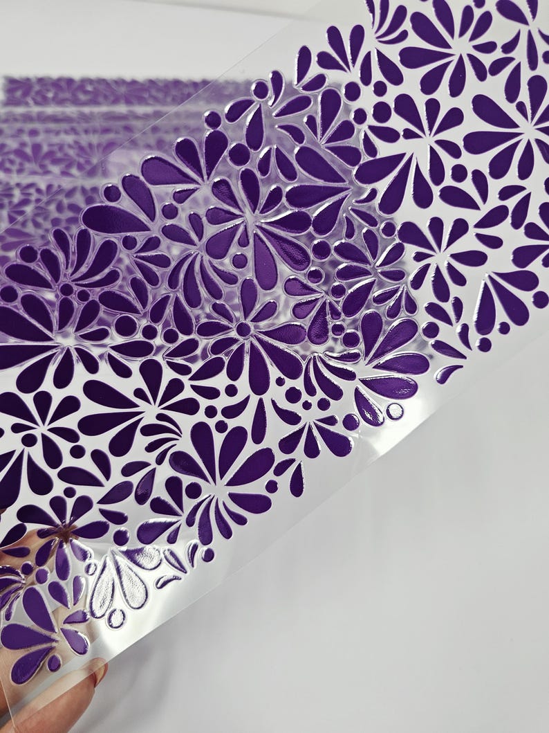 May include: A sheet of clear plastic with a repeating pattern of purple floral designs. The floral pattern is embossed, creating a raised texture. The design is a mix of stylized flowers and teardrop shapes.