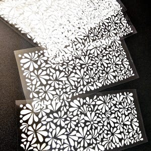 May include: Several clear plastic sheets with a white floral pattern. The design features stylized white flowers and leaf shapes. The sheets are stacked, with the top one partially obscuring the others. The background is black.