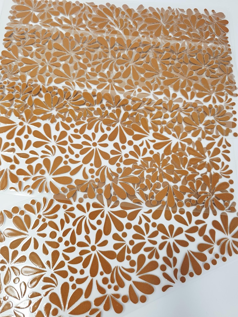 May include: Close-up of a sheet of brown floral stickers with a clear backing. The stickers feature a repeating pattern of stylized flowers and teardrop shapes. The stickers are arranged in rows and are partially peeled away from the backing.