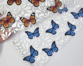 May include: A clear adhesive sheet featuring multiple butterfly stickers in orange and black or blue and black patterns. The background of the sheet is decorated with a repeating white leaf-like pattern, and the butterflies are arranged in a scattered layout.