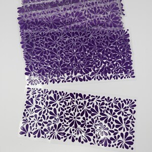 May include: Several sheets of clear plastic with a repeating pattern of purple floral designs. The floral pattern is composed of teardrop and circular shapes. The sheets are stacked and arranged on a white background.