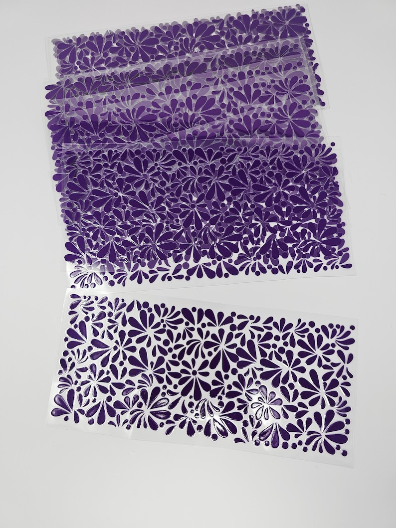May include: Several sheets of clear plastic with a repeating pattern of purple floral designs. The floral pattern is composed of teardrop and circular shapes. The sheets are stacked and arranged on a white background.