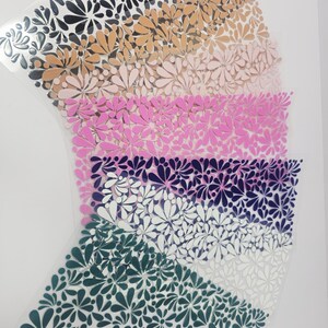 May include: Sheets of floral-patterned vinyl in various colors, including black, tan, pink, purple, white, and teal. The floral design is a repeating pattern of stylized flowers. The vinyl sheets are arranged in a layered, fan-like shape.