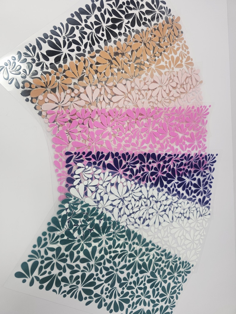 May include: Sheets of floral-patterned vinyl in various colors, including black, tan, pink, purple, white, and teal. The floral design is a repeating pattern of stylized flowers. The vinyl sheets are arranged in a layered, fan-like shape.