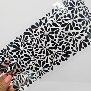 May include: A clear, rectangular stencil featuring a repeating pattern of black floral designs. The floral elements are stylized with teardrop and dot shapes. The stencil is held against a white background.