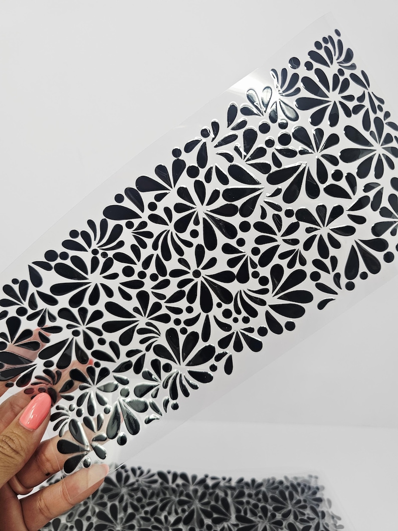 May include: A clear, rectangular stencil featuring a repeating pattern of black floral designs. The floral elements are stylized with teardrop and dot shapes. The stencil is held against a white background.