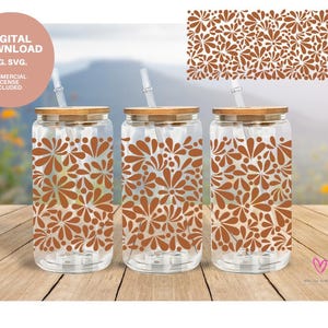 May include: Three clear glass tumblers with brown floral patterns and bamboo lids. Each tumbler has a straw inserted through the lid. The tumblers are on a wooden surface with a blurred background of green foliage.