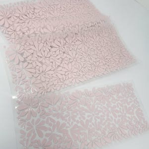 May include: Two sheets of pink floral patterned material. The sheets are transparent and the floral design is raised. The flowers are a soft pink color and cover the entire surface of the sheets. The sheets are laying on a white surface.