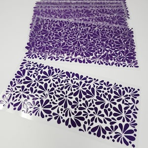 May include: Several sheets of clear, transparent material are stacked, each adorned with a repeating pattern of dark purple floral designs. The floral pattern is composed of stylized petals and circular shapes, creating a decorative border.