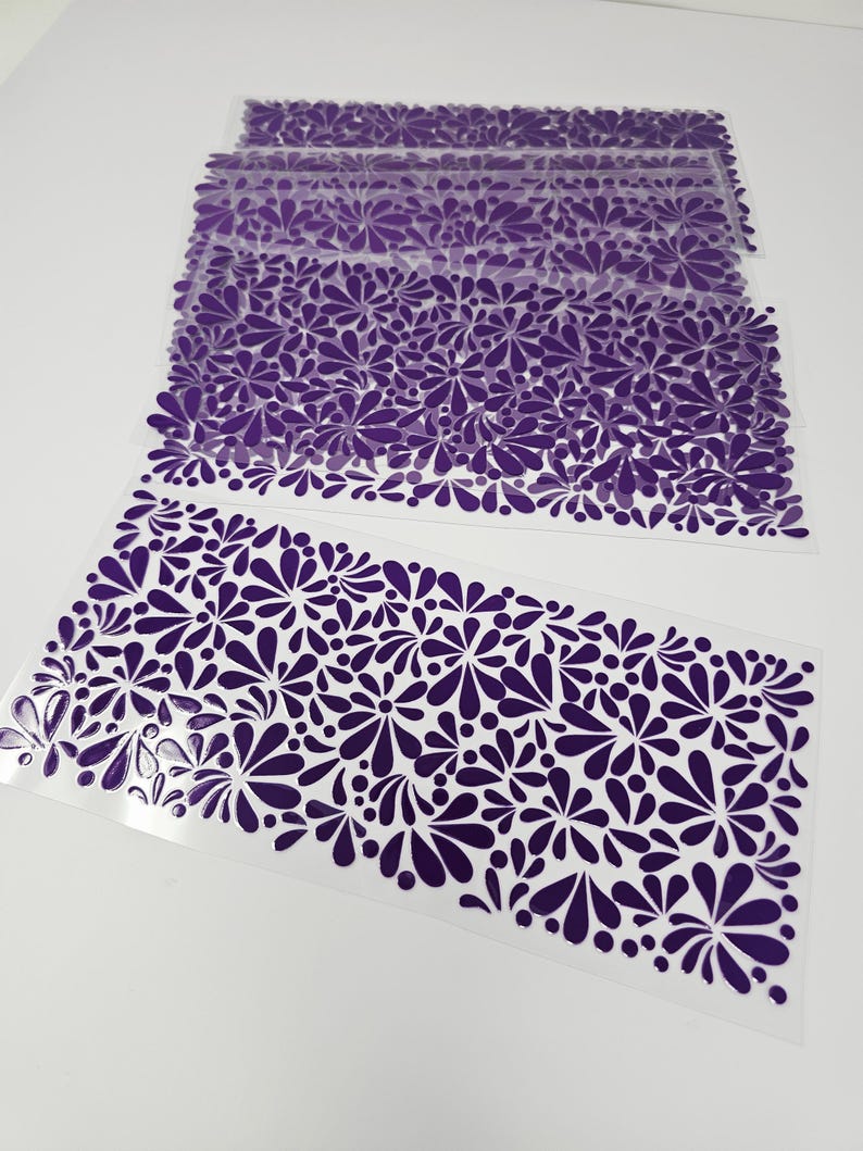 May include: Several sheets of clear, transparent material are stacked, each adorned with a repeating pattern of dark purple floral designs. The floral pattern is composed of stylized petals and circular shapes, creating a decorative border.