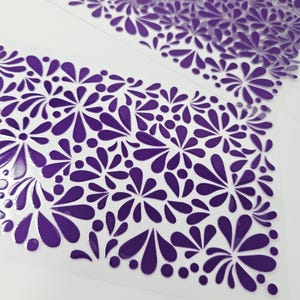 May include: Close-up of a clear sheet with a repeating pattern of purple floral designs. The design features stylized flowers and teardrop shapes, creating a decorative and textured surface. The pattern is evenly spaced across the sheet.