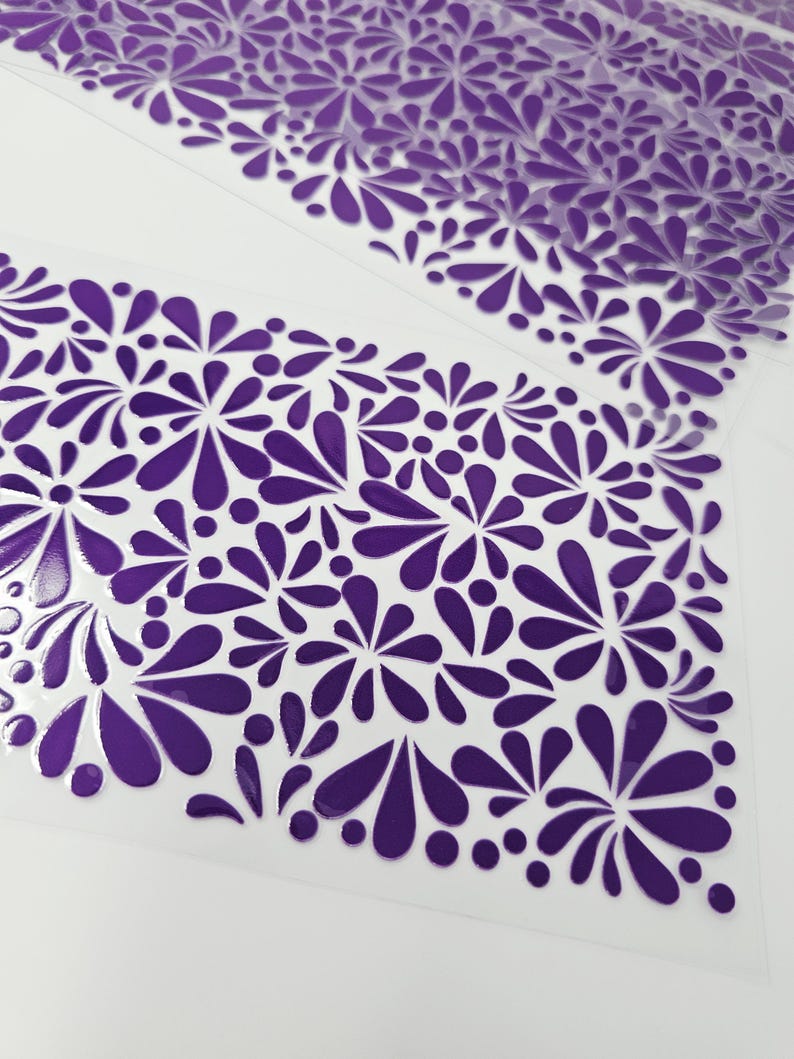 May include: Close-up of a clear sheet with a repeating pattern of purple floral designs. The design features stylized flowers and teardrop shapes, creating a decorative and textured surface. The pattern is evenly spaced across the sheet.