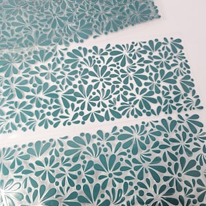 May include: Close-up of several sheets of teal-colored floral patterned material. The floral design features a repeating pattern of stylized flowers and teardrop shapes. The sheets are transparent and appear to be made of plastic or a similar material.