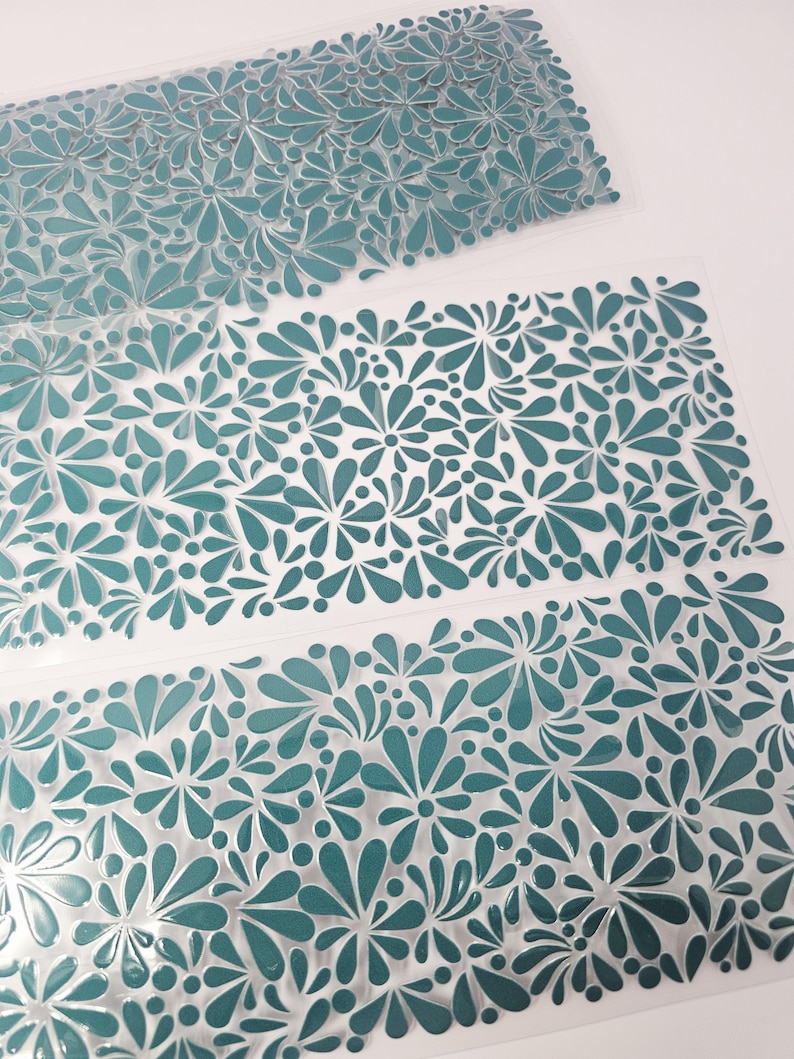 May include: Close-up of several sheets of teal-colored floral patterned material. The floral design features a repeating pattern of stylized flowers and teardrop shapes. The sheets are transparent and appear to be made of plastic or a similar material.