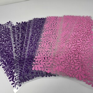 May include: A stack of clear plastic sheets with a floral design. The sheets transition from deep purple to pink. The floral pattern is made up of teardrop-shaped petals, creating a textured appearance. The sheets are stacked on a white surface.