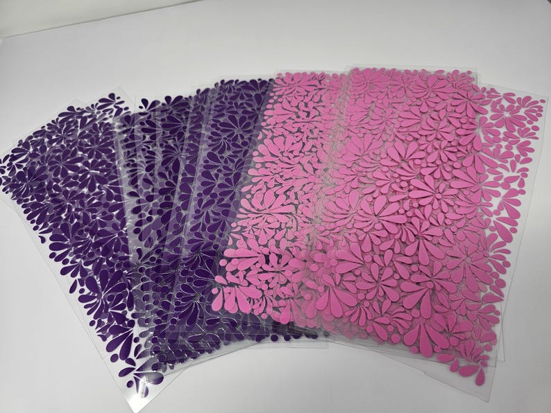 May include: A stack of clear plastic sheets with a floral design. The sheets transition from deep purple to pink. The floral pattern is made up of teardrop-shaped petals, creating a textured appearance. The sheets are stacked on a white surface.