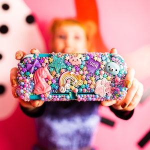 May include: A teal Nintendo Switch game console with a custom case covered in colorful rhinestones, glitter, and charms. The case features a unicorn, rainbow, stars, and the word "UGH".