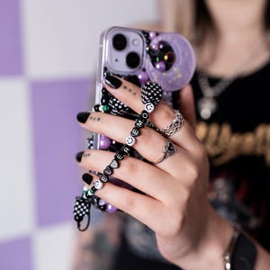 Può includere: Una custodia per telefono in bianco e nero con un retro trasparente e un cinturino per telefono con perline in bianco e nero con la parola "ELDER" scritta in perline nere. La custodia del telefono ha un retro trasparente con un design scintillante e un anello per telefono a scacchi in bianco e nero.