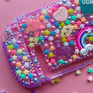 Puede incluir: Funda de tel&eacute;fono morada decorada con estrellas, perlas y abalorios multicolores. La funda presenta un coraz&oacute;n rosa con "XOXO" y una gota azul con "UGH". Tambi&eacute;n se incluye un arco&iacute;ris, una nube y un unicornio.