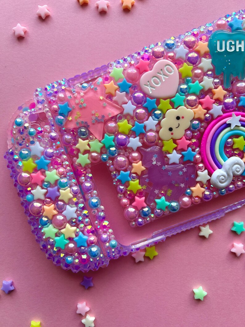 Puede incluir: Funda de tel&eacute;fono morada decorada con estrellas, perlas y abalorios multicolores. La funda presenta un coraz&oacute;n rosa con "XOXO" y una gota azul con "UGH". Tambi&eacute;n se incluye un arco&iacute;ris, una nube y un unicornio.