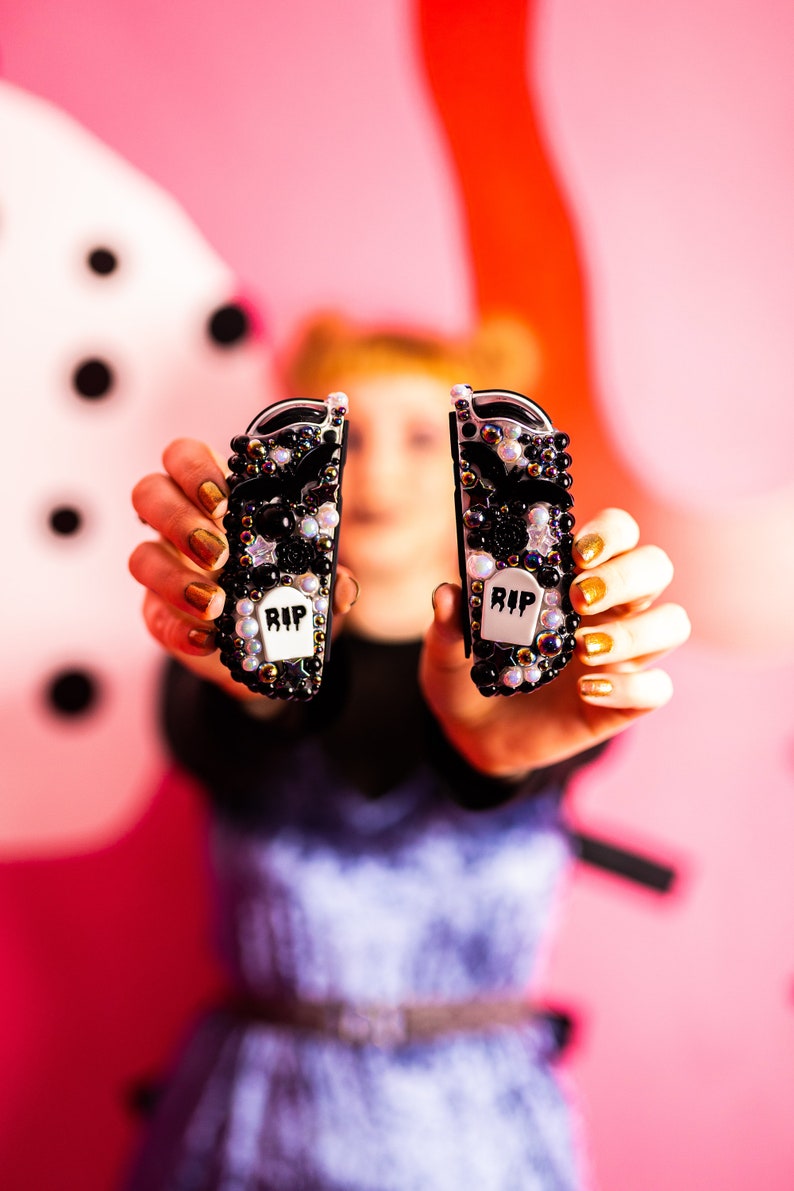 May include: Two black and white decorated Nintendo Switch Joy-Con controllers with a "RIP" tombstone design. The controllers are held up in front of a pink background.