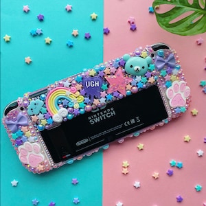 May include: A pink and blue Nintendo Switch case decorated with stars, a rainbow, a cloud, a paw print, a bear, and the word "UGH".
