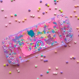 Puede incluir: Una funda transparente para Nintendo Switch llena de confeti, estrellas y abalorios de colores. La funda presenta un coraz&oacute;n rosa, un arco&iacute;ris y una luna creciente. Los mandos est&aacute;n adornados con cuentas iridiscentes, todo sobre un fondo rosa.