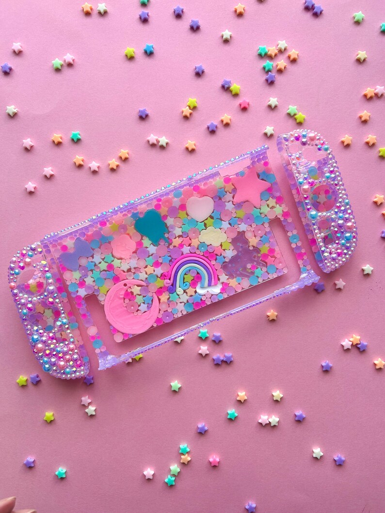 Puede incluir: Una funda transparente para Nintendo Switch llena de confeti, estrellas y abalorios de colores. La funda presenta un coraz&oacute;n rosa, un arco&iacute;ris y una luna creciente. Los mandos est&aacute;n adornados con cuentas iridiscentes, todo sobre un fondo rosa.