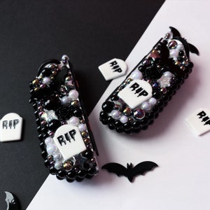 May include: Two black and white decorated game controllers with a Halloween theme. The controllers are covered in black beads, white pearls, and small white tombstones with the word "RIP" on them. There are also small black bats and stars on the controllers.