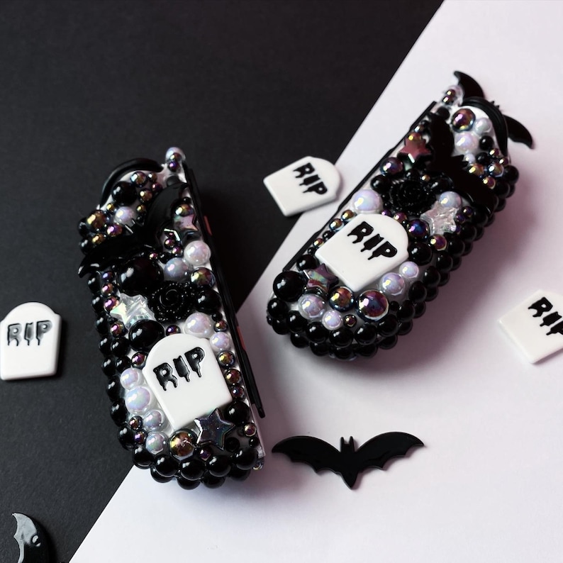 May include: Two black and white decorated game controllers with a Halloween theme. The controllers are covered in black beads, white pearls, and small white tombstones with the word "RIP" on them. There are also small black bats and stars on the controllers.