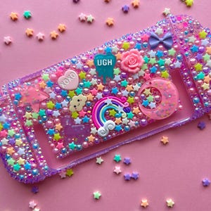 Puede incluir: Una funda decorativa morada y transparente para un dispositivo de juego, adornada con cuentas en forma de estrella de colores, perlas y abalorios. La funda presenta un arco&iacute;ris, una luna creciente, una rosa y la palabra "UGH". El fondo es rosa liso.