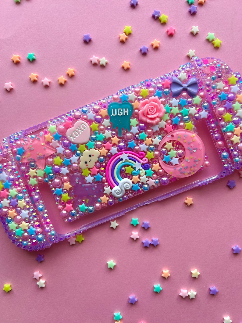 Puede incluir: Una funda decorativa morada y transparente para un dispositivo de juego, adornada con cuentas en forma de estrella de colores, perlas y abalorios. La funda presenta un arco&iacute;ris, una luna creciente, una rosa y la palabra "UGH". El fondo es rosa liso.