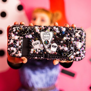 May include: A black phone case covered in iridescent rhinestones, pearls, and charms. The charms include a black and white "RIP" charm, a black and white "UGH" charm, and a black and white Ouija board charm.