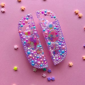 Puede incluir: Dos mandos Joy-Con Switch morados y transparentes, adornados con cuentas iridiscentes, perlas y confeti en forma de estrella. Los mandos est&aacute;n sobre un fondo rosa salpicado de decoraciones en forma de estrella.