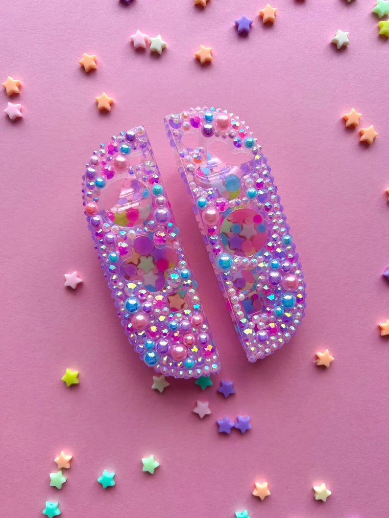 Puede incluir: Dos mandos Joy-Con Switch morados y transparentes, adornados con cuentas iridiscentes, perlas y confeti en forma de estrella. Los mandos est&aacute;n sobre un fondo rosa salpicado de decoraciones en forma de estrella.