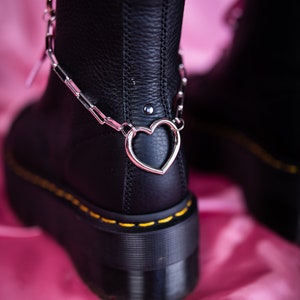 Könnte beinhalten: Schwarze Lederstiefel mit einer dicken Plateau-Sohle und einer silbernen Kette mit einem Herz-Anhänger um den Knöchel.