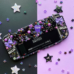 Puede incluir: Funda negra para consola Nintendo Switch decorada con cuentas, estrellas y un lazo morados, negros e iridiscentes. La funda tiene las palabras "OUJIA" y "UGH". El fondo está dividido entre negro y morado.