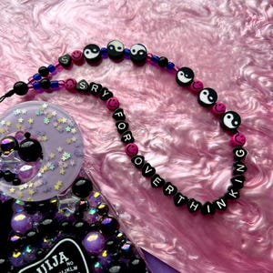 May include: A black beaded necklace with pink and white beads spelling out "Sorry for overthinking" with yin yang symbols and smiley faces. The necklace is on a pink and white marbled surface.